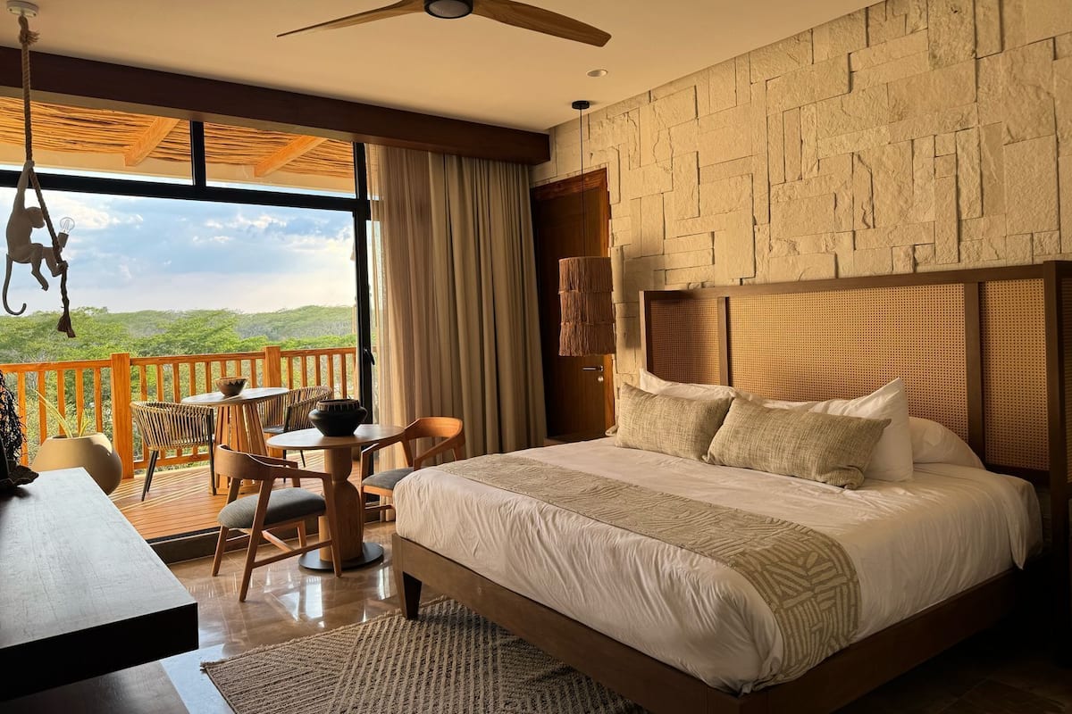 A bedroom with a large bed, a wooden headboard, a dining table with chairs, and a view of the outdoors.