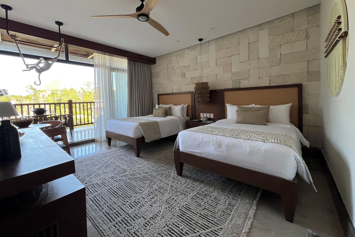 A hotel room with two beds, a ceiling fan, a dining area with a table and chairs, and a view of the outdoors.