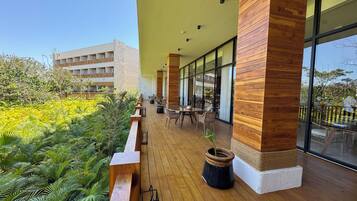 Un espace extérieur abrité, doté d’un plancher en bois, de sièges et de grandes fenêtres offrant une vue sur un bâtiment et une végétation luxuriante.