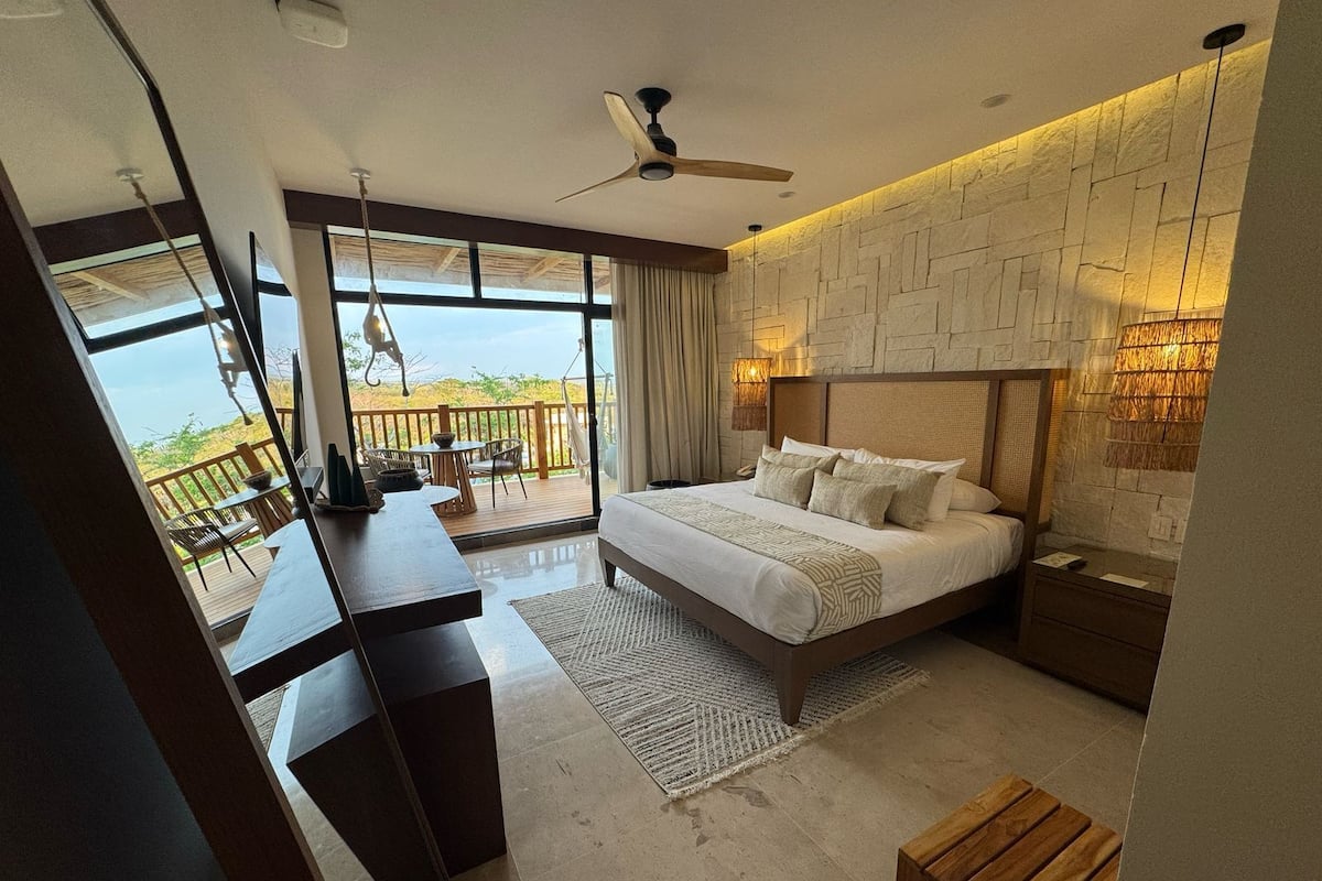 A bedroom with a large bed, a desk, a chair, a balcony with a view, and a ceiling fan.