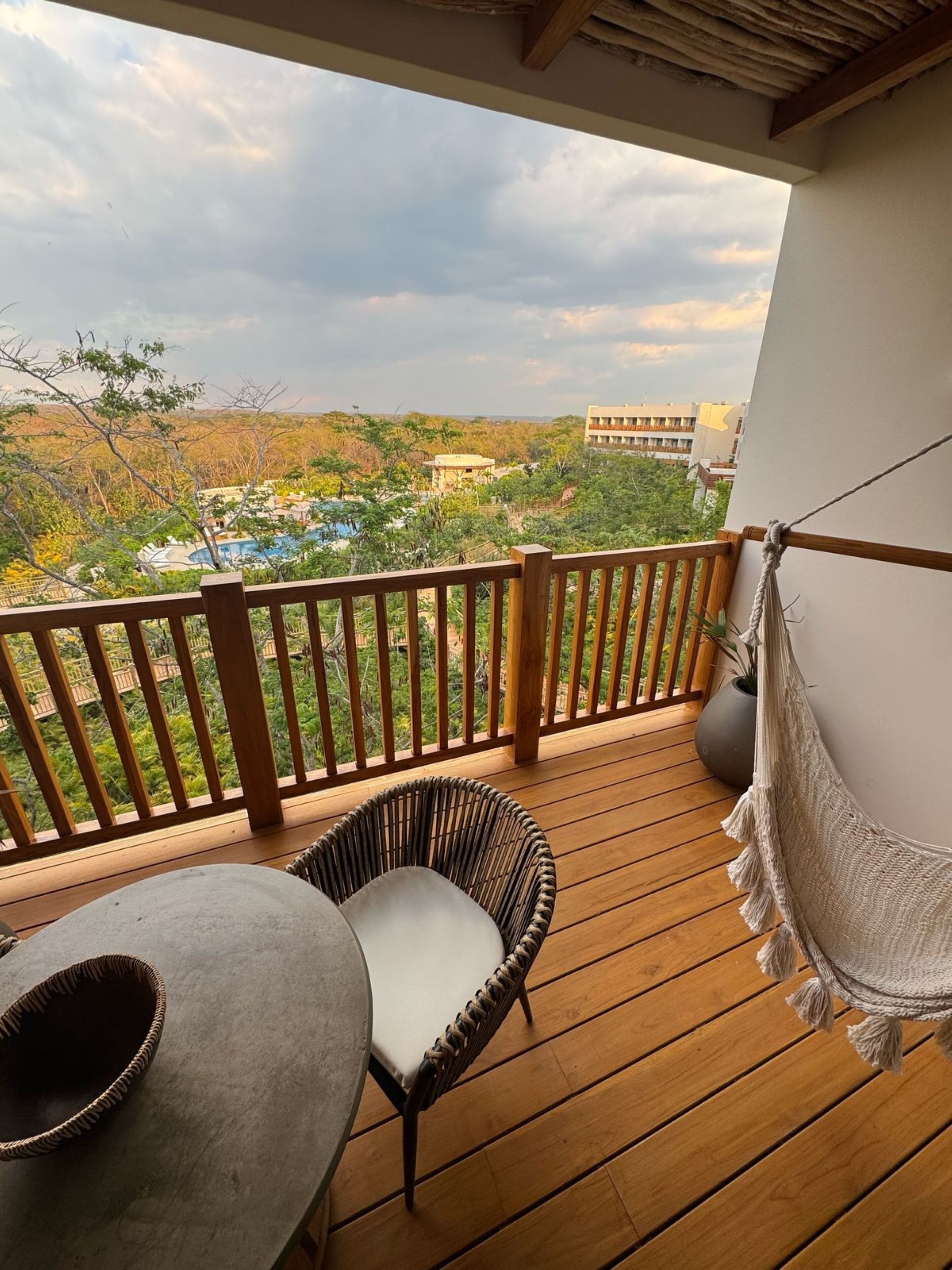 Une terrasse en bois avec une chaise en osier et une table, d’où l’on peut admirer un paysage luxuriant avec des bâtiments visibles au loin.
