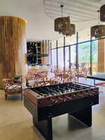 Un espacio interior moderno con mesa de fútbol de mesa, sillas de mimbre y una zona de bar con un ventanal que ofrece vistas al exterior.