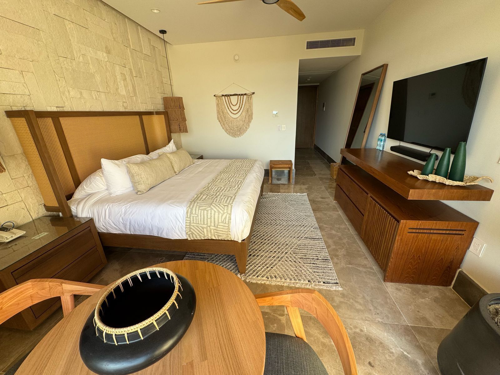 A modern hotel room with a large bed, a wooden headboard, a TV, a small table, and a seating area with a woven chair.