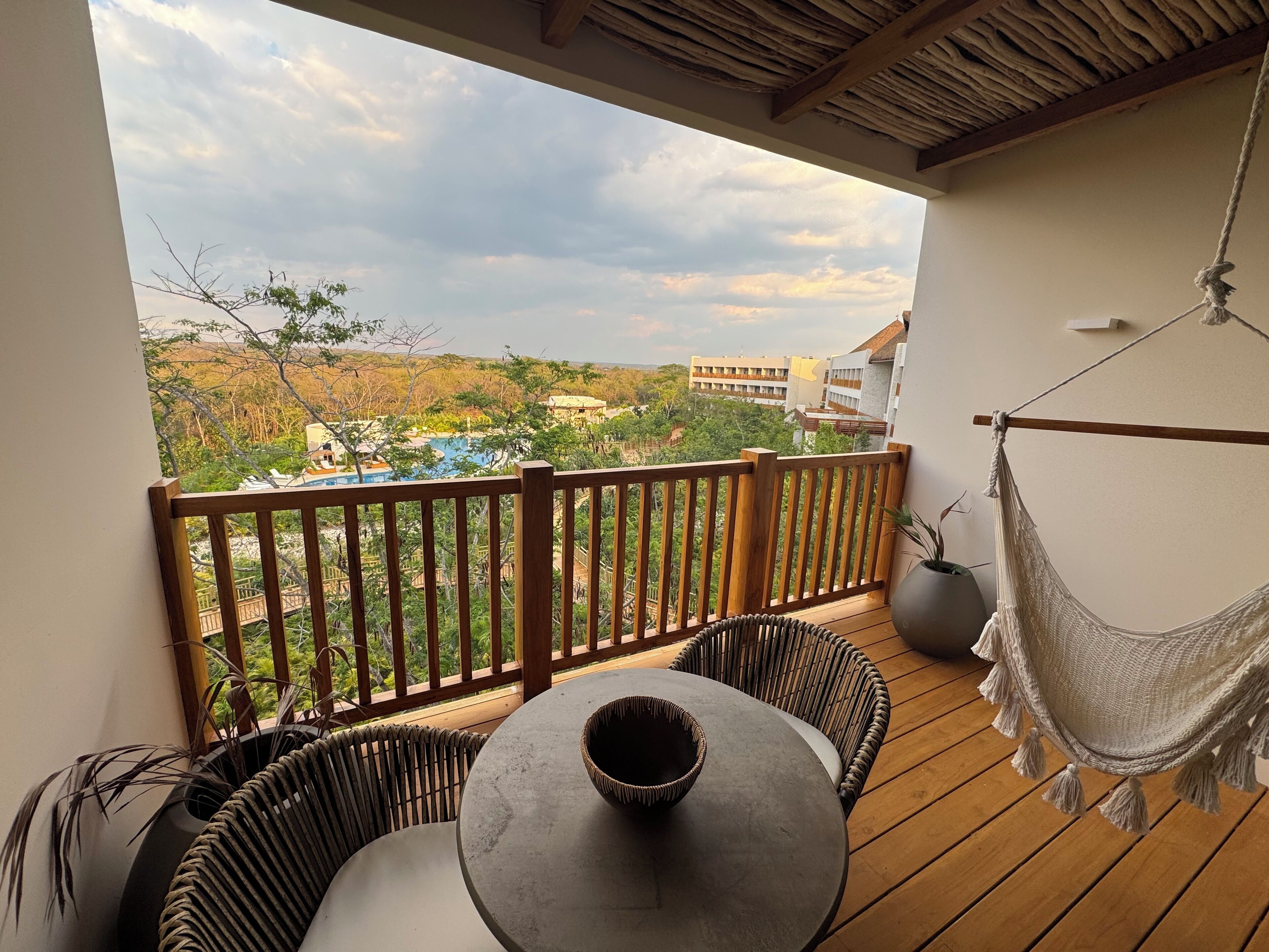 Une terrasse en bois avec une table ronde et des chaises, un hamac, et une vue sur une piscine et des bâtiments au loin.