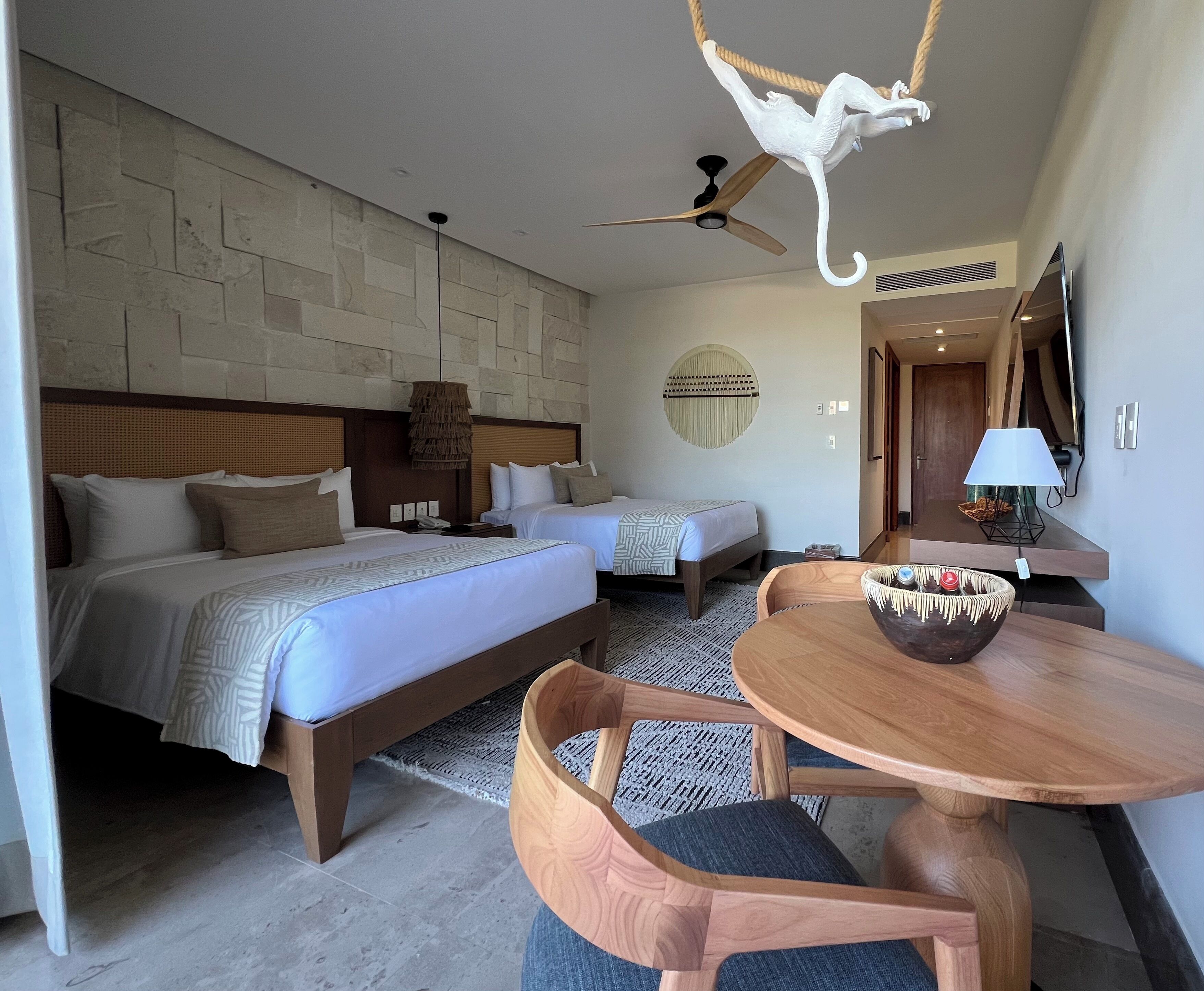 Quarto de hotel com duas camas, uma mesa de jantar, um ventilador de teto e uma escultura única de macaco.