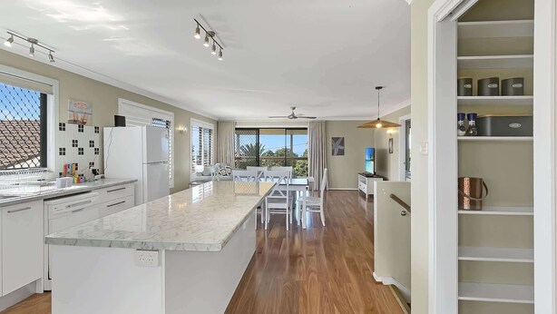 A modern kitchen with a marble island, wooden flooring, and a dining area with white furniture. - Spacious 4-Bed Coastal Home with Water Views (Batehaven)