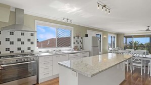 A modern kitchen with a central island, stainless steel appliances, and large windows. - Spacious 4-Bed Coastal Home with Water Views (Batehaven)