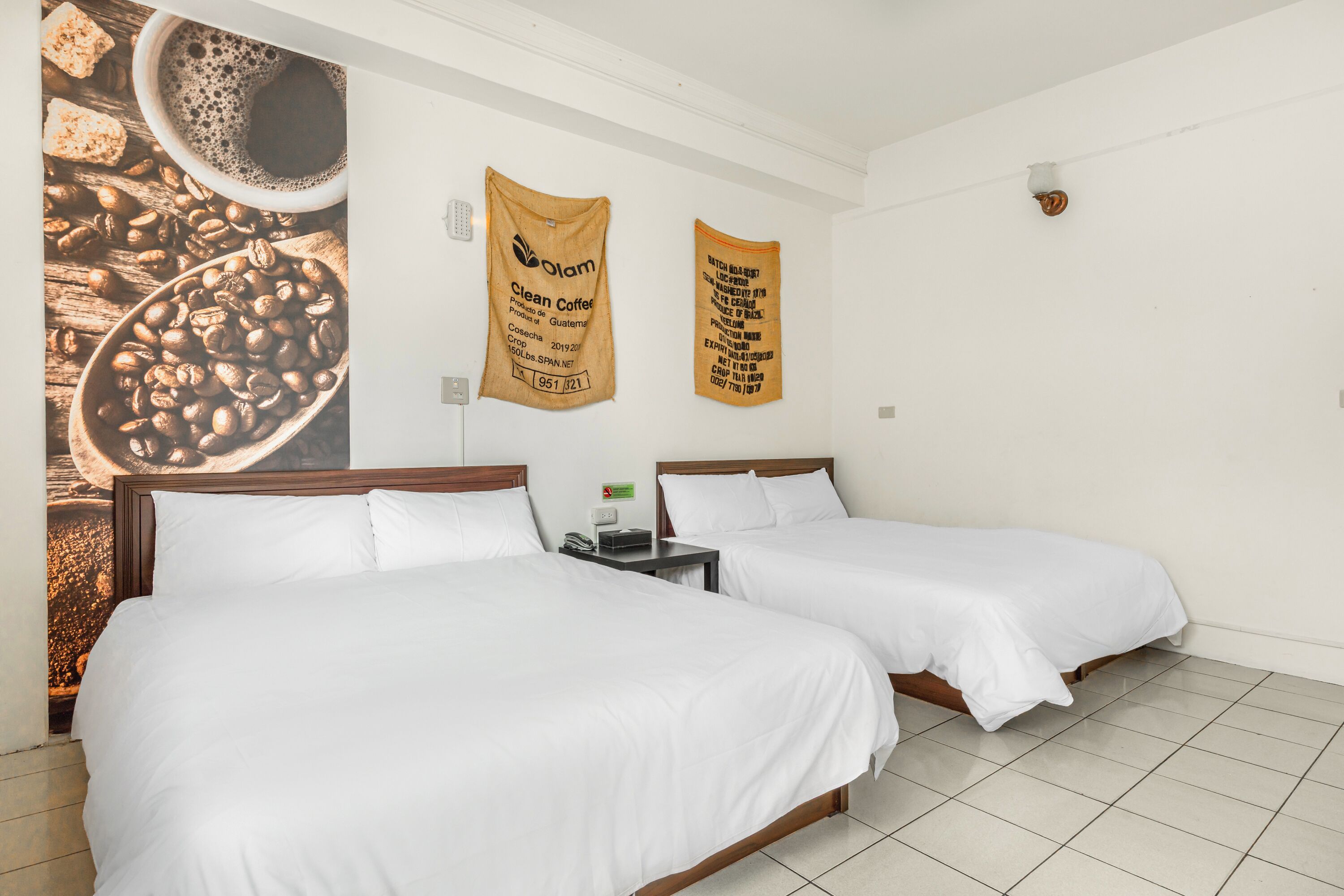 Una habitació d'hotel amb dos llits, una obra d'art mural amb tema de cafè i dos banners publicitaris de 'Clean Coffee'.