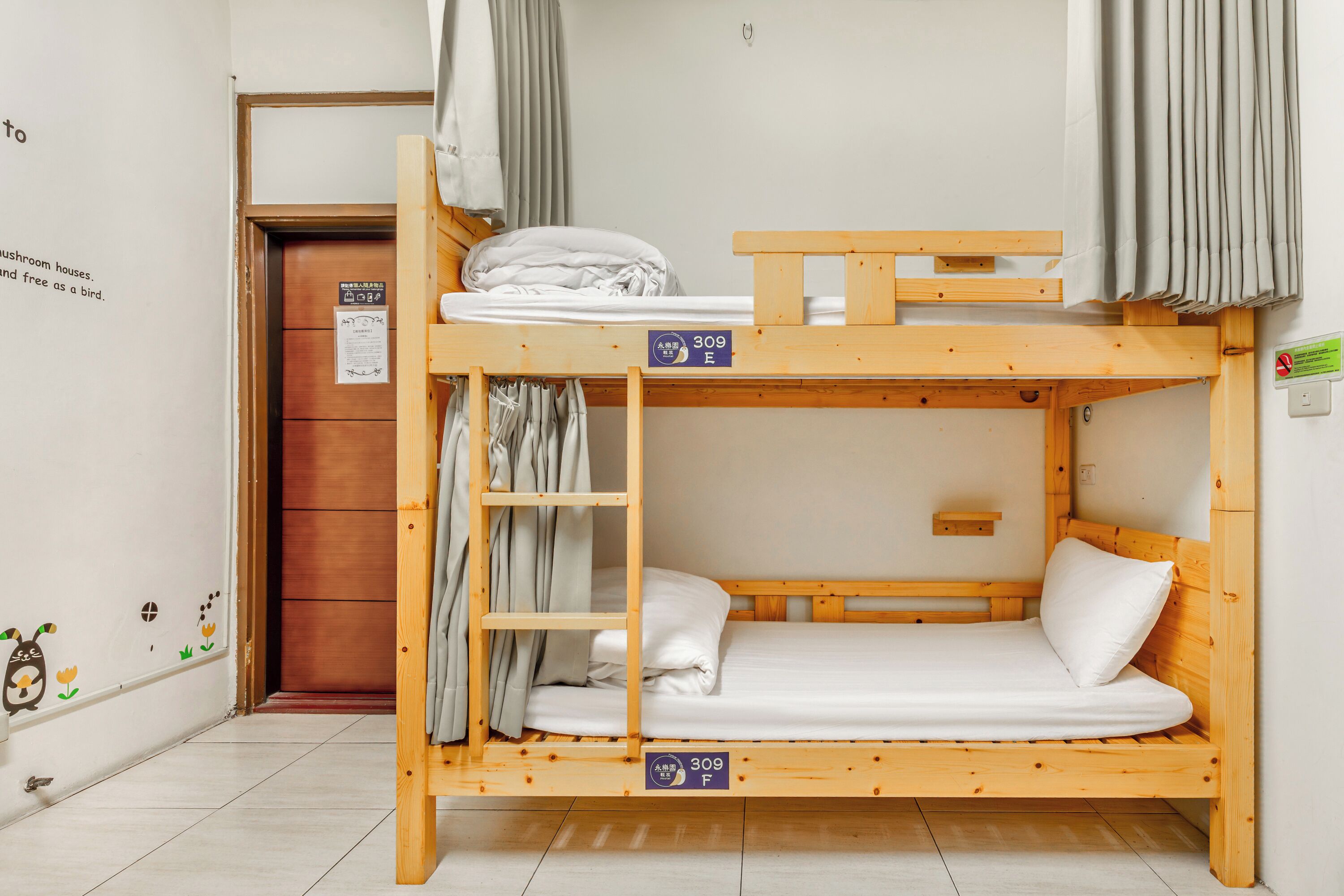 A bunk bed with a wooden frame and a mattress. There is a curtain on the upper bunk and a door with a sign on the lower bunk.