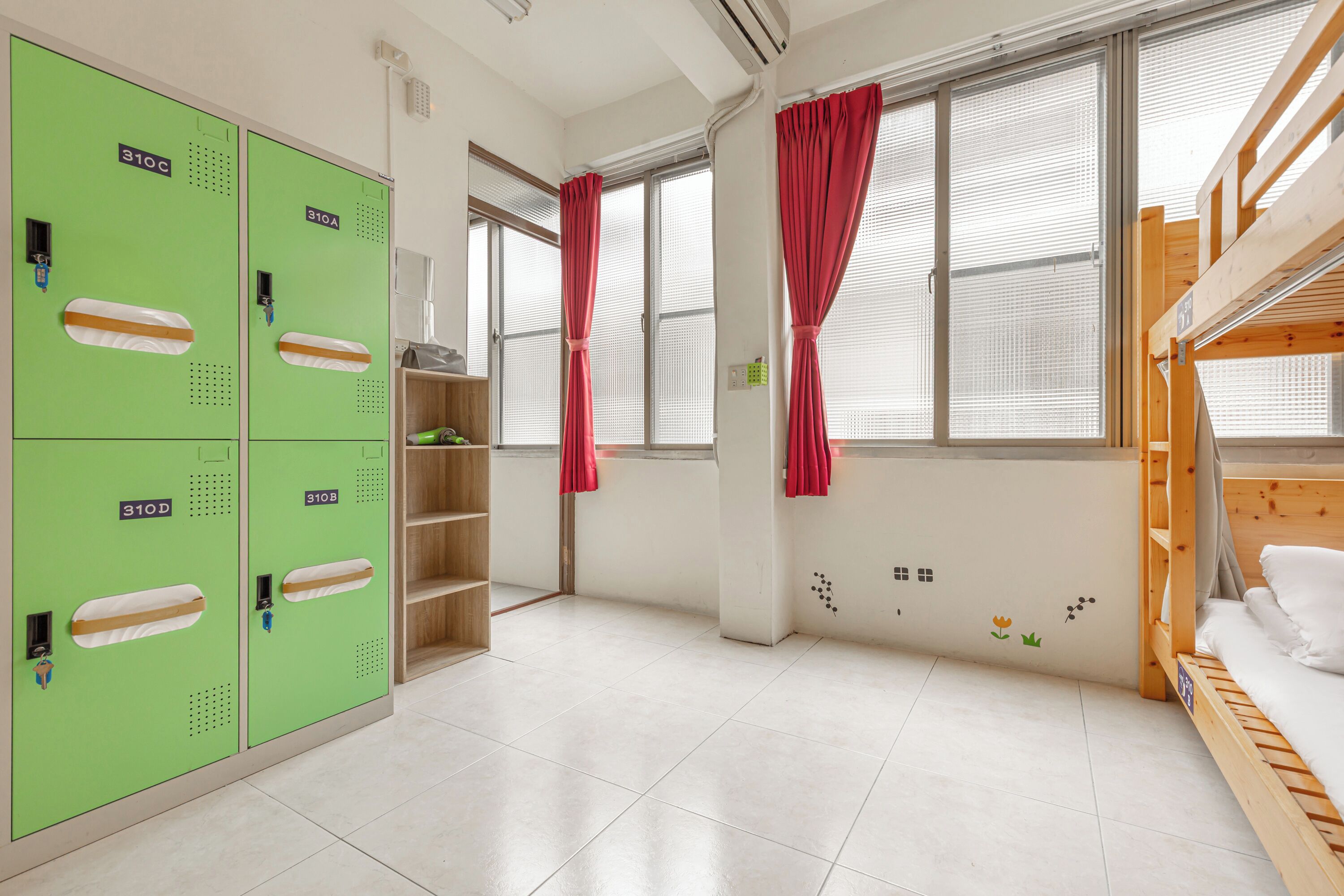 A room with bunk beds, green lockers, and a window with red curtains.