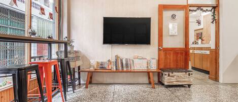 A room with a large window, a flat-screen TV, a wooden bench, and a small table with books.