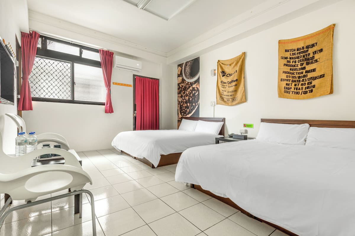 Ein Hotelzimmer mit zwei Betten, einem Schreibtisch und einem Stuhl. Rote Vorhänge sind an den Fenstern und an der Wand hängt ein Banner mit Kaffeebohnen.