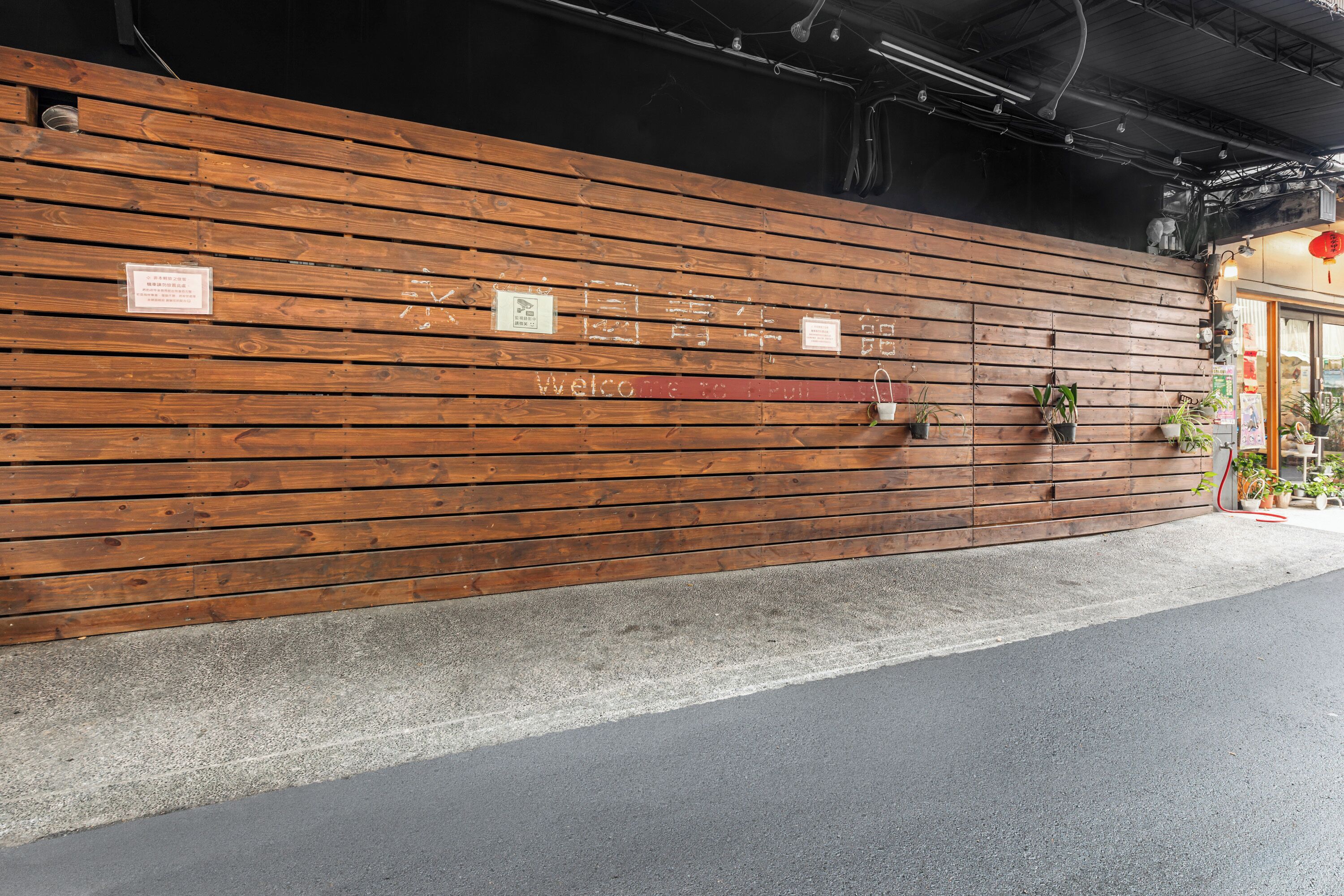 A wooden wall with horizontal planks, a welcome sign, and several potted plants.
