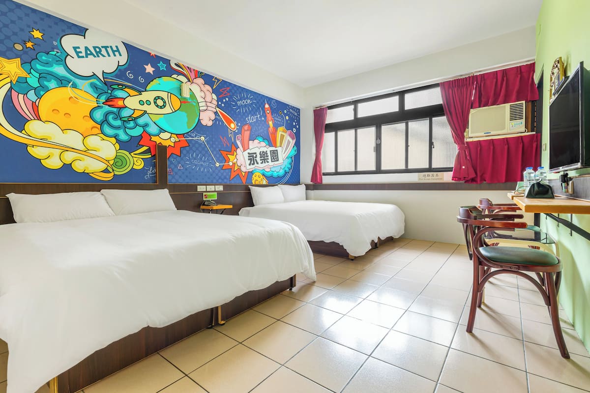 Ein Hotelzimmer mit zwei Betten, einem Wandgemälde mit Weltraumkunst, einem Fenster mit roten Vorhängen und einem Schreibtisch mit Stuhl.