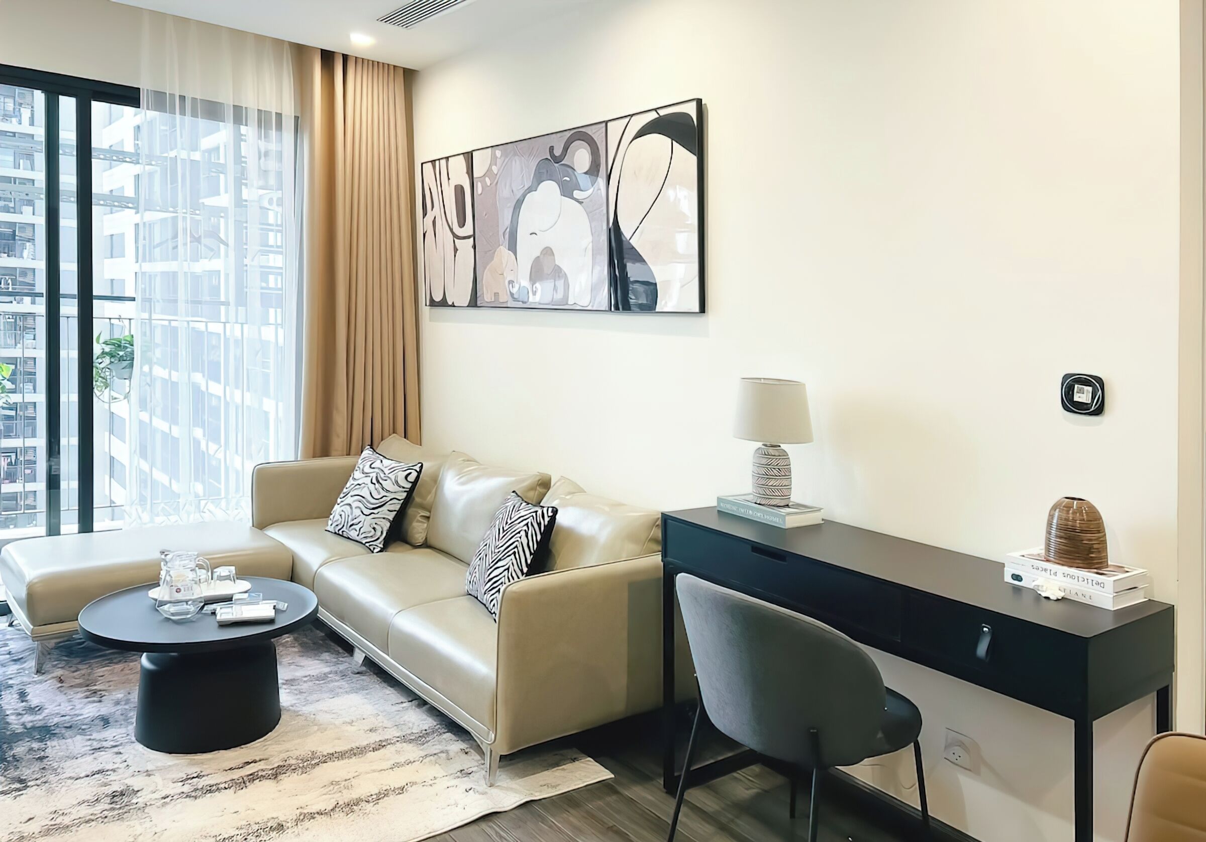 Una sala de estar moderna con un sofá, una mesa de centro, un escritorio y una silla. Hay un ventanal con cortinas, un cuadro en la pared y una lámpara sobre el escritorio.