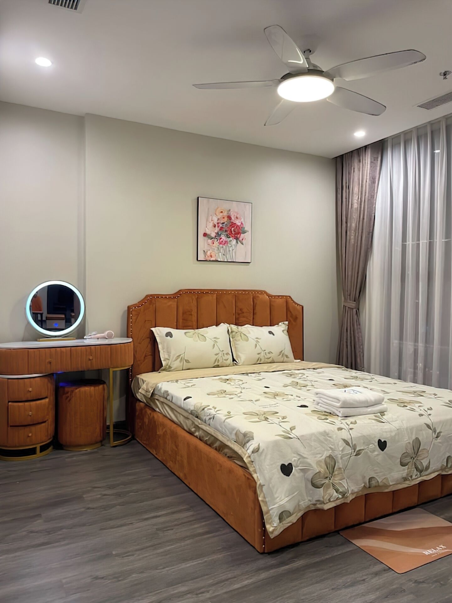 A bedroom with a wooden bed, a bedside table with a mirror, a ceiling fan, and a floral painting on the wall.