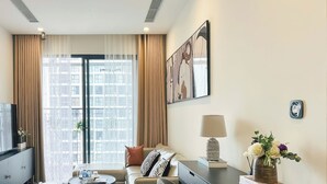 A modern living room with a sofa, coffee table, and a TV on a cabinet. There is a large window with curtains, a painting on the wall, and a lamp on a side table. - The Zenpark Tower - Vinhomes Ocean Park (Hanoi)