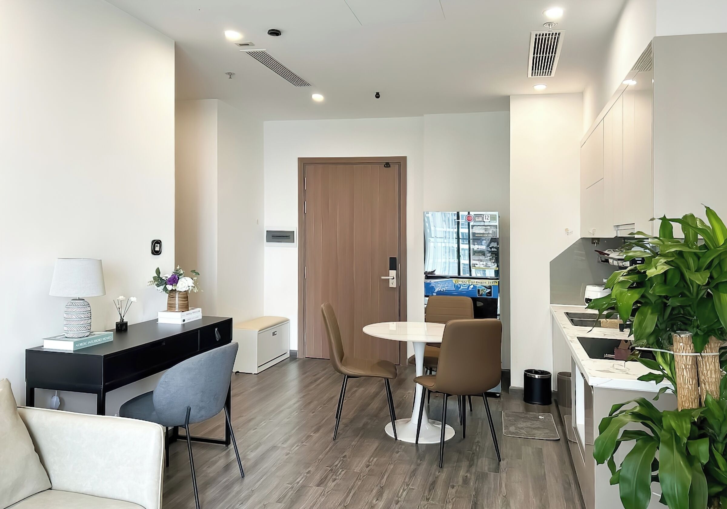 A modern hotel room with a desk, two chairs, a small table, a sofa, a kitchen area, and a door leading to another room.