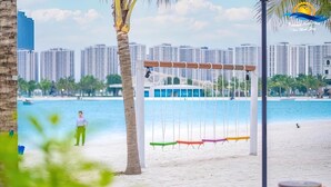 A beach with a swing set, palm trees, and a cityscape in the background. - The Zenpark Tower - Vinhomes Ocean Park (Hanoi)