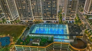 A high-rise building with a large glass-covered swimming pool, surrounded by greenery and adjacent to a road. - The Zenpark Tower - Vinhomes Ocean Park (Hanoi)