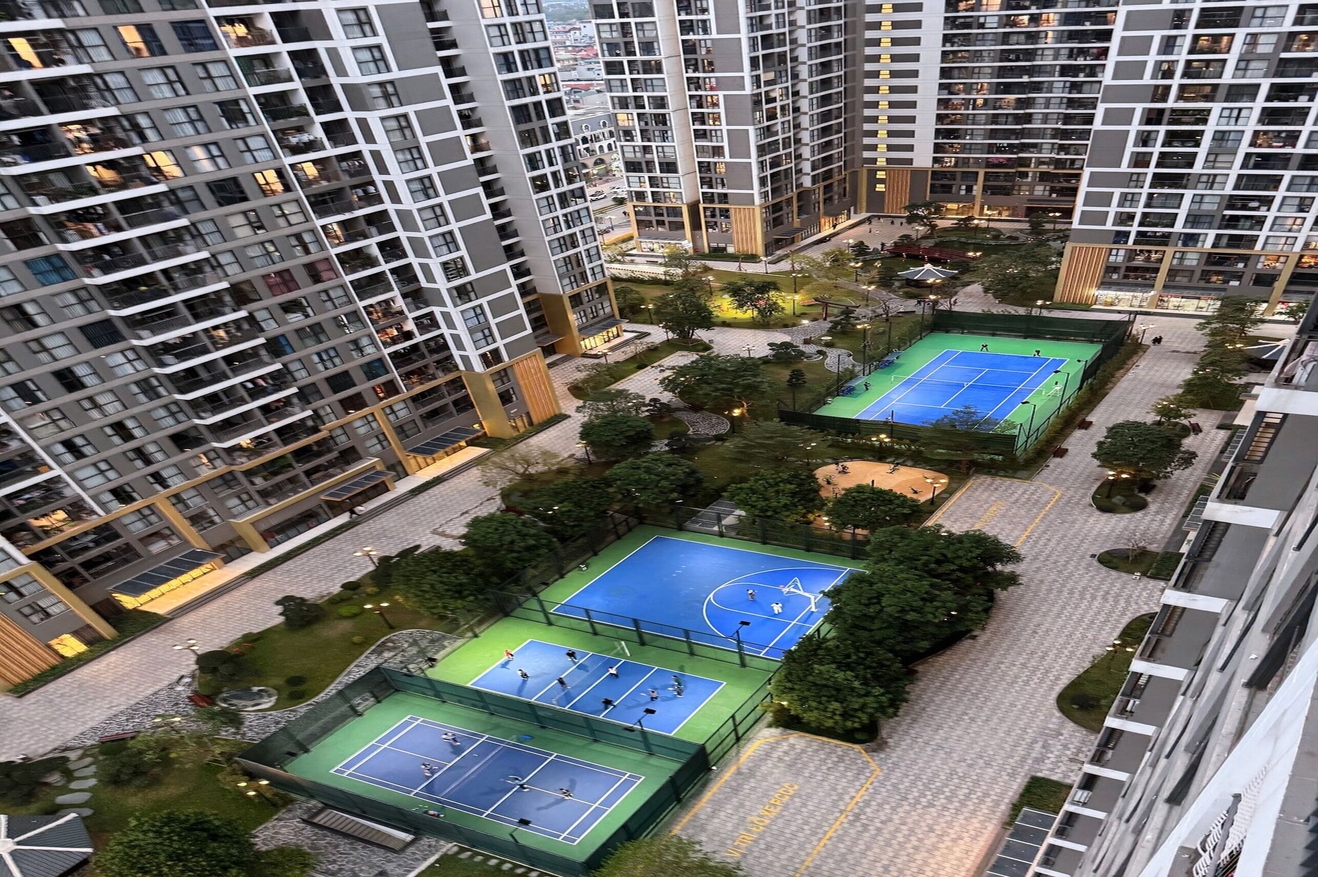 Un edificio residencial de gran altura con varios balcones, un patio central con canchas de tenis y zonas verdes circundantes.