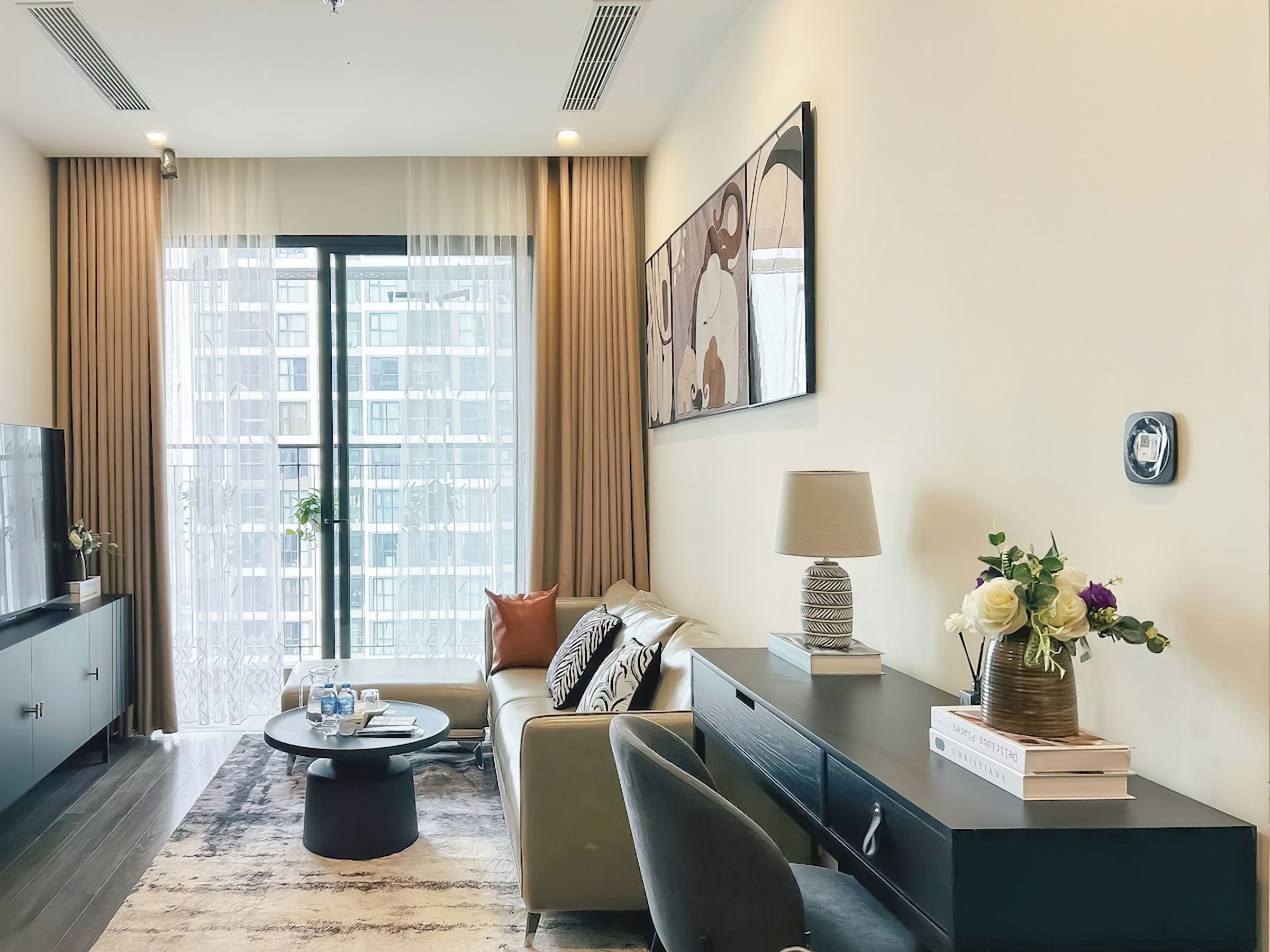 Uma sala de estar moderna com sofá, mesa de centro e televisão em um armário. Há uma janela grande com cortinas, um quadro na parede e um abajur em uma mesinha.