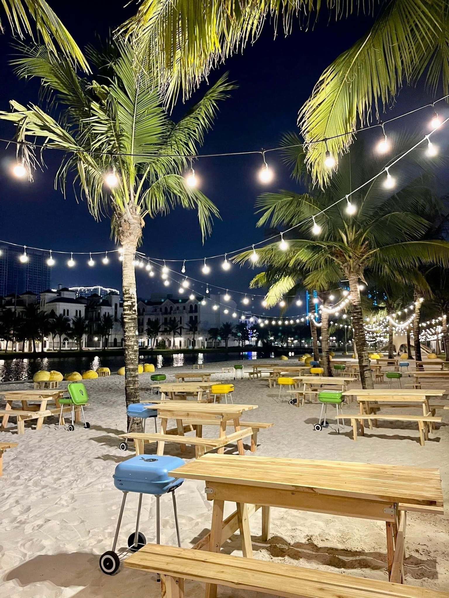 A beachside area with wooden picnic tables and benches, palm trees, and string lights.