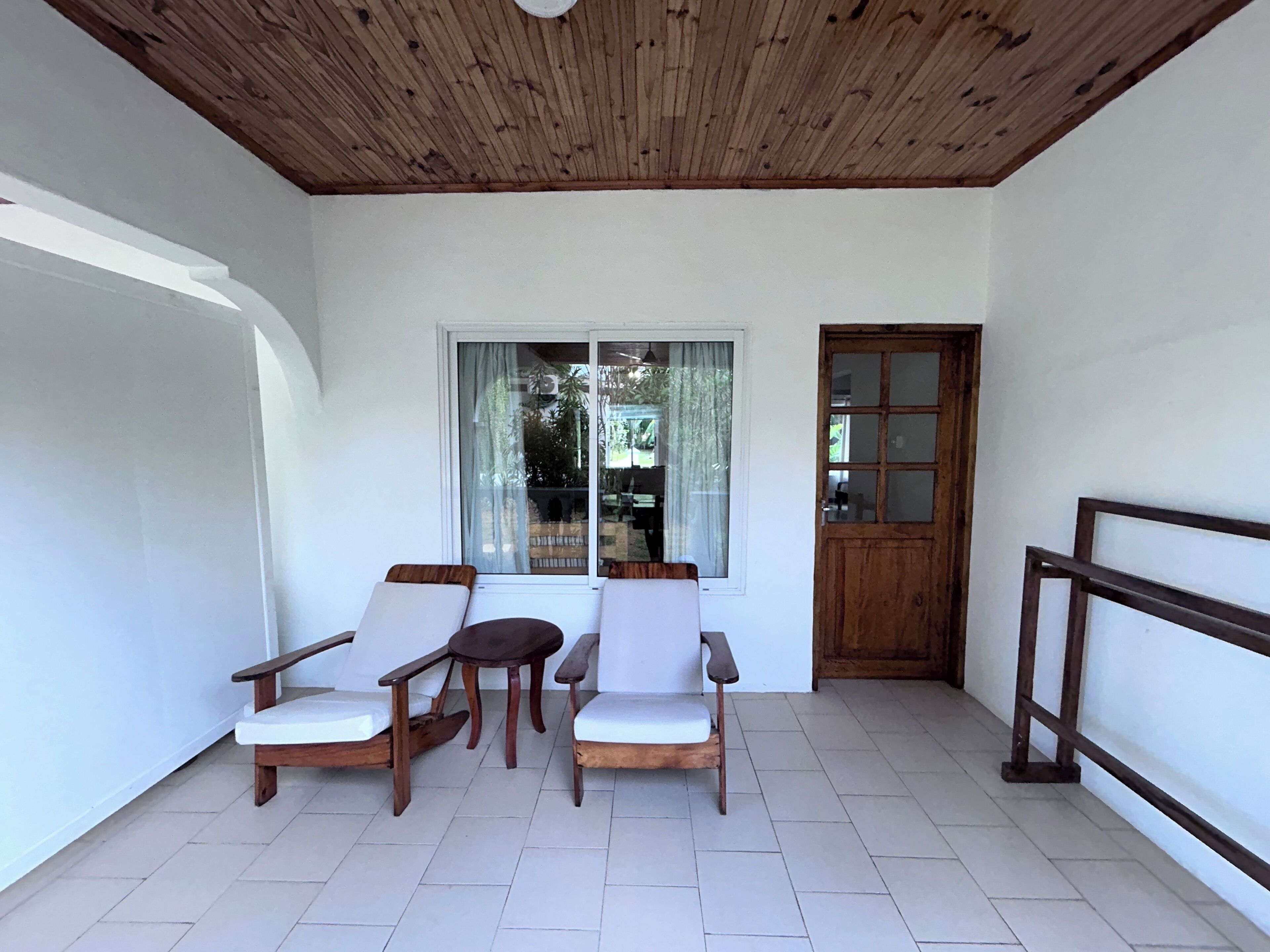 Habitación con techo de madera, paredes blancas y piso de baldosas. Hay dos sillas de madera con cojines blancos, una pequeña mesa de madera y una ventana con vista a la vegetación.