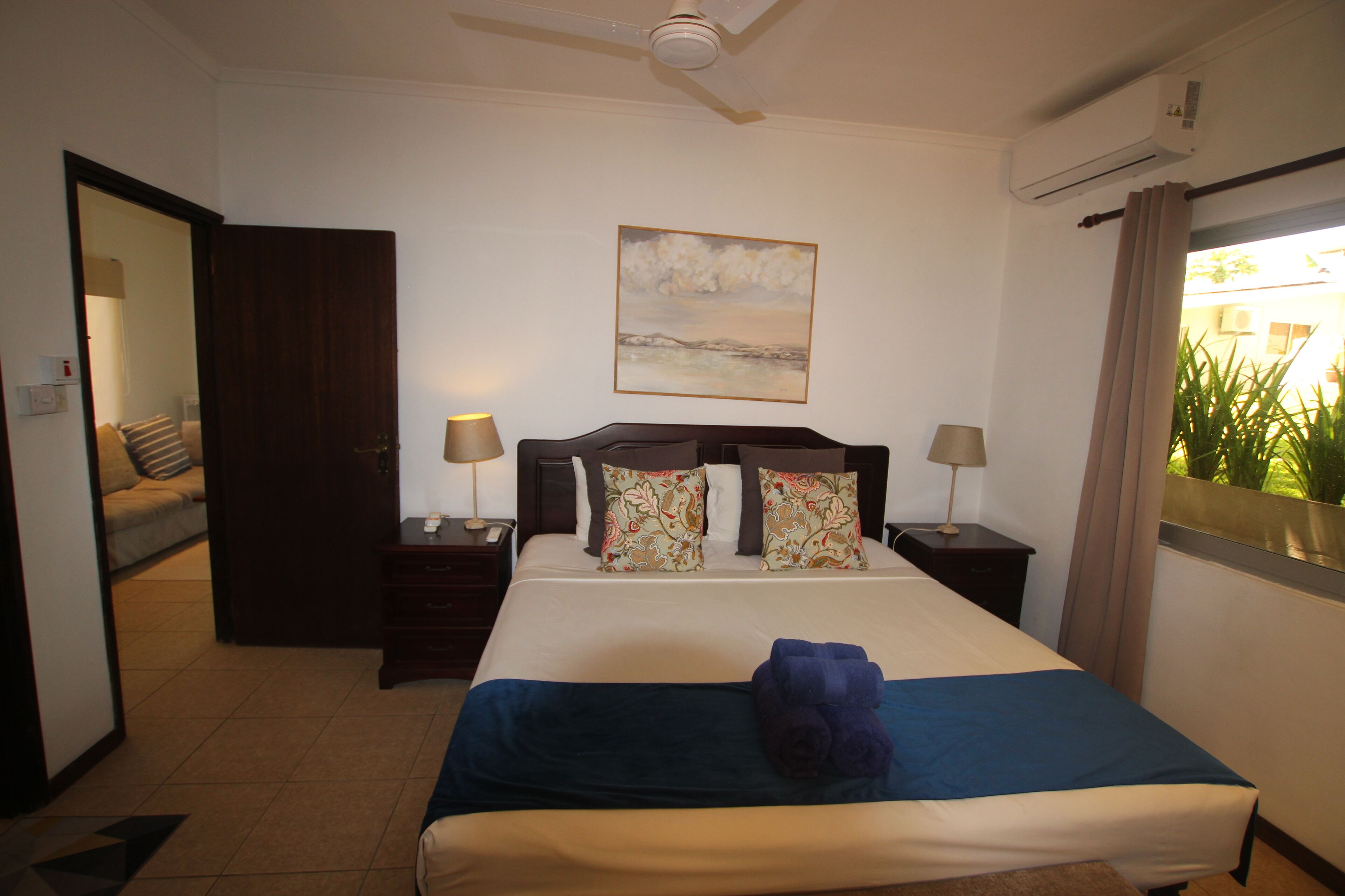 A hotel room with a bed, two bedside tables with lamps, a ceiling fan, an air conditioning unit, and a window with curtains.