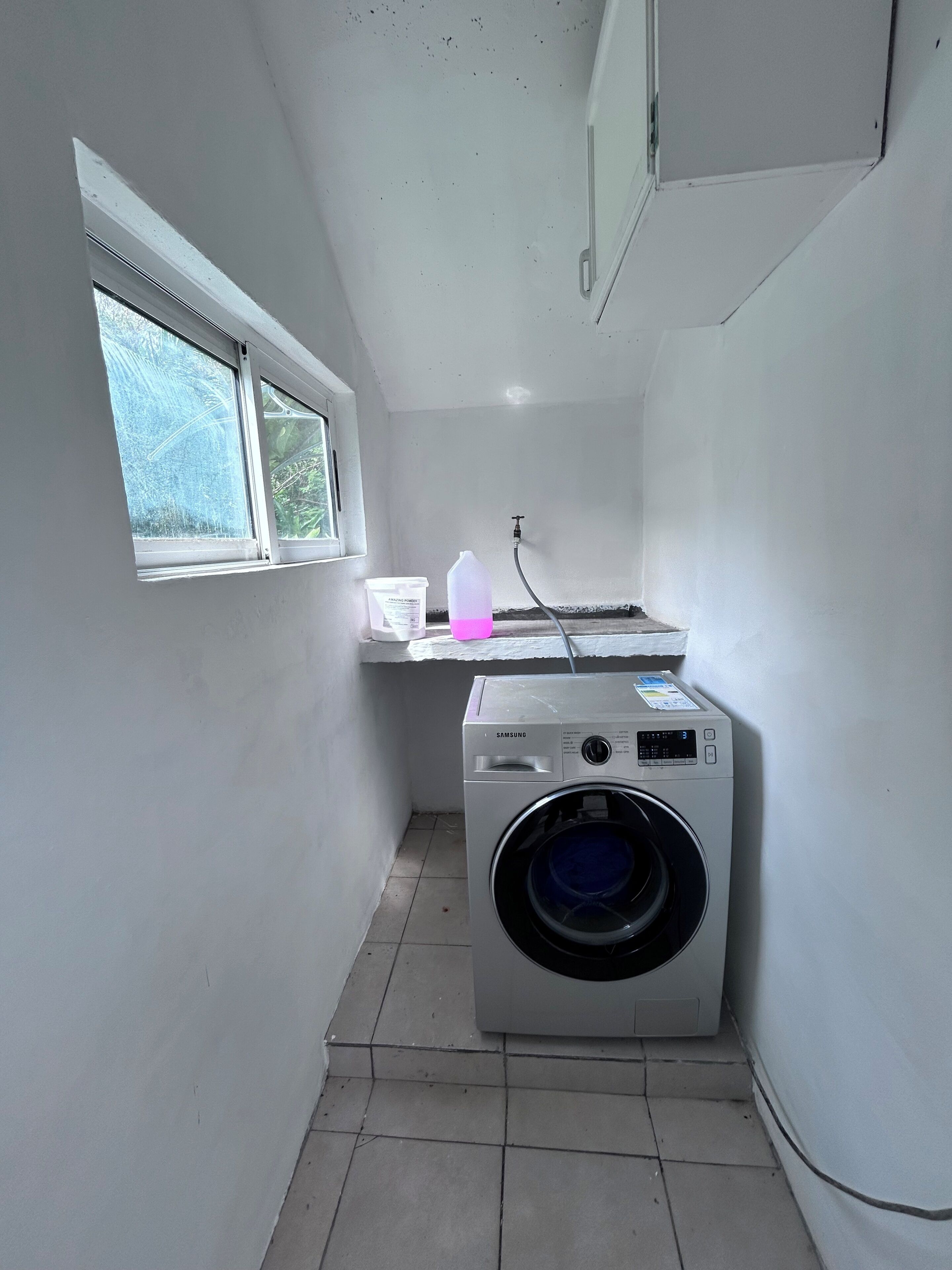 A laundry room with a Samsung washing machine, a window, and a small countertop with a water bottle and a container.