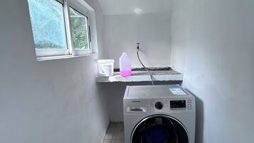 A laundry room with a Samsung washing machine, a window, and a small countertop with a water bottle and a container.