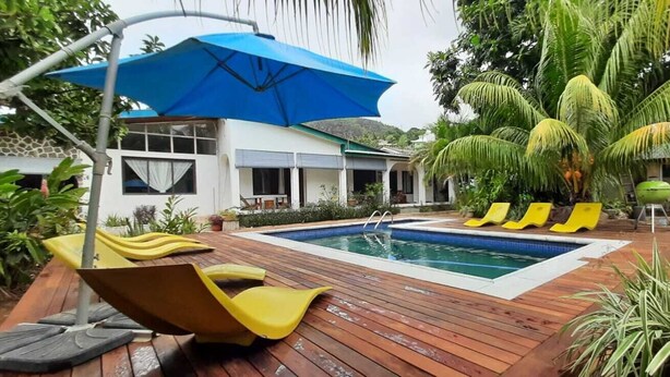 A swimming pool with wooden deck and yellow lounge chairs, surrounded by tropical plants and a blue umbrella. - castaway lodge (Pointe Au Sel)