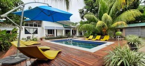 A swimming pool with wooden deck and yellow lounge chairs, surrounded by tropical plants and a blue umbrella. - castaway lodge (Pointe Au Sel)