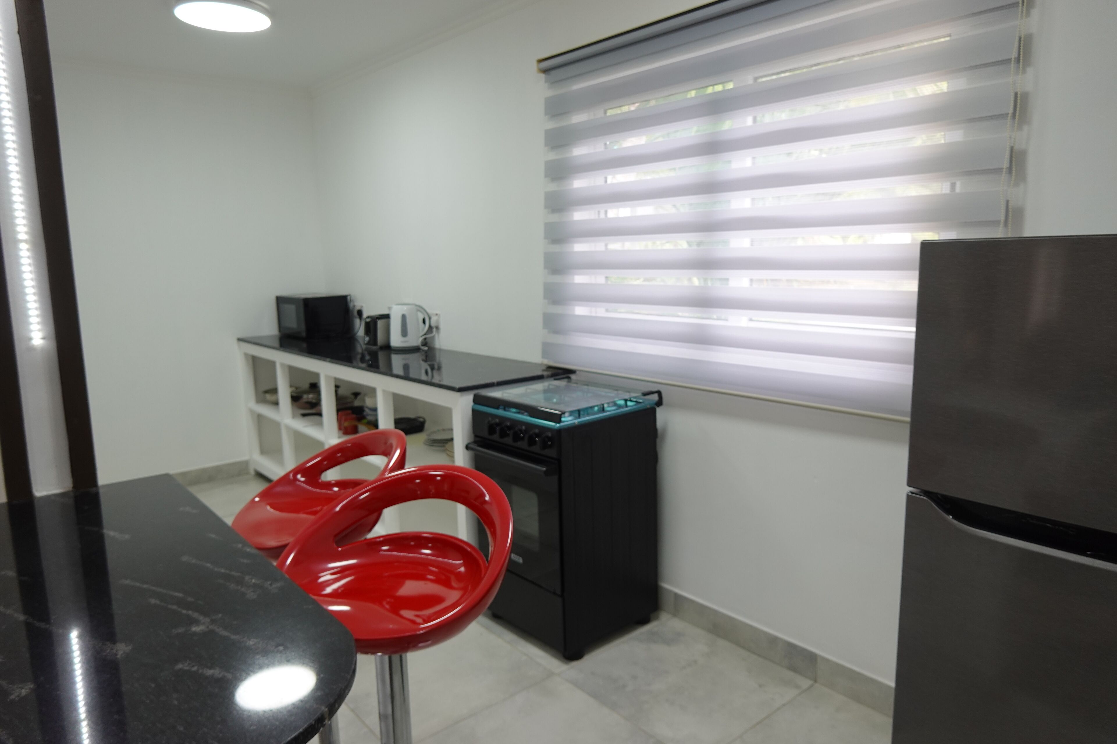 A compact kitchen with a black countertop, a red chair, a stove, and a refrigerator.
