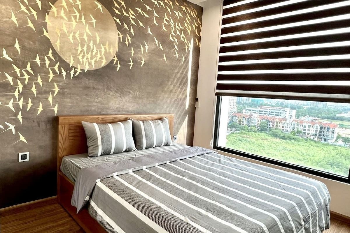 A bedroom with a large bed, a view of buildings through the window, and a wall with a bird design.