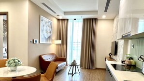 A modern hotel room with a dining area, a sofa, a small table with a lamp, and a kitchen area with a sink and countertop. - Vinhomes Green Bay Apartments (Hanoi)