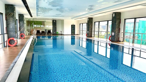 A large indoor swimming pool with clear blue water, surrounded by walls with marble-like patterns, and large windows offering views of the outside. - Vinhomes Green Bay Apartments (Hanoi)
