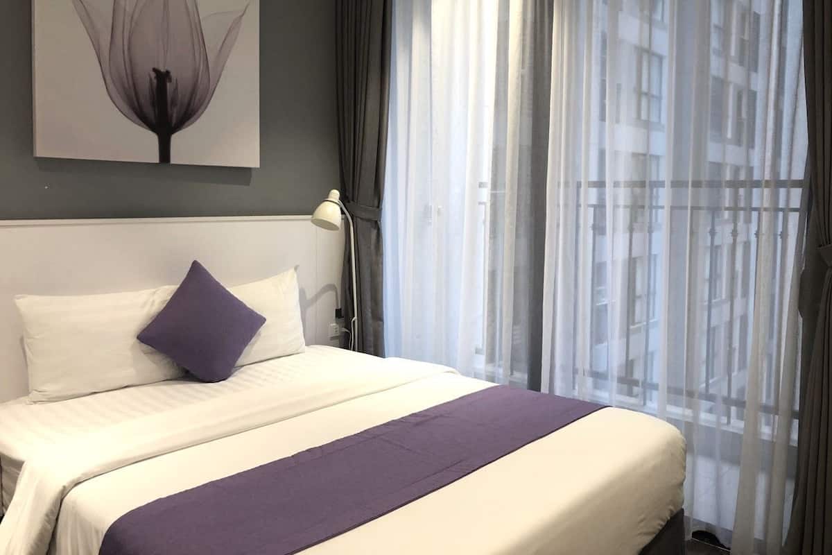 A neatly made bed with white and purple bedding, a single purple pillow, and a wall-mounted lamp.