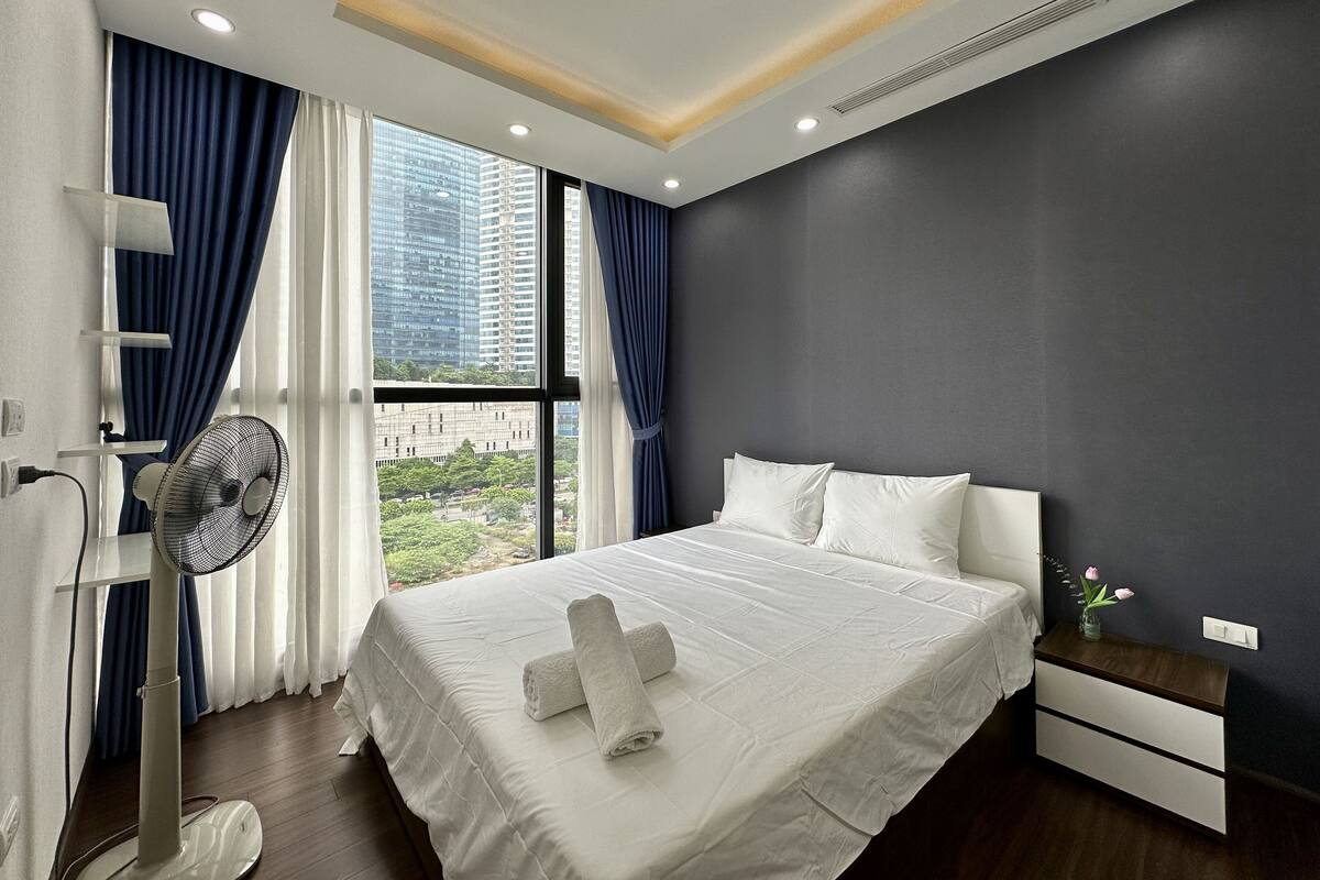 A modern hotel room with a large bed, a bedside table, a nightstand, a fan, and a view of the city through large windows.