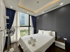 Een moderne hotelkamer met een groot bed, nachtkastje, ventilator en uitzicht op de stad door grote ramen.