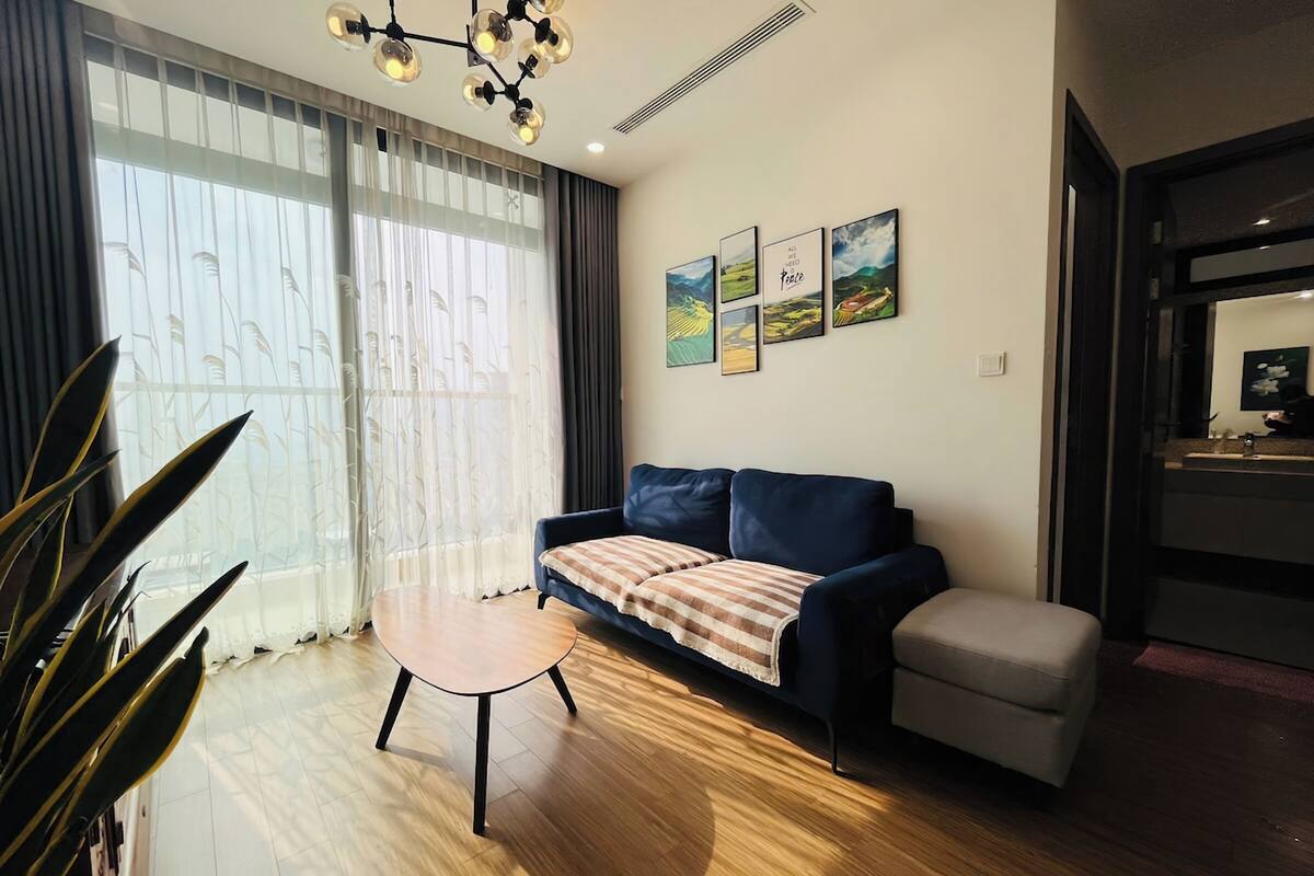 A modern living room with a sofa, a small table, and a bench. There are large windows with sheer curtains, a chandelier, and framed artwork on the wall.