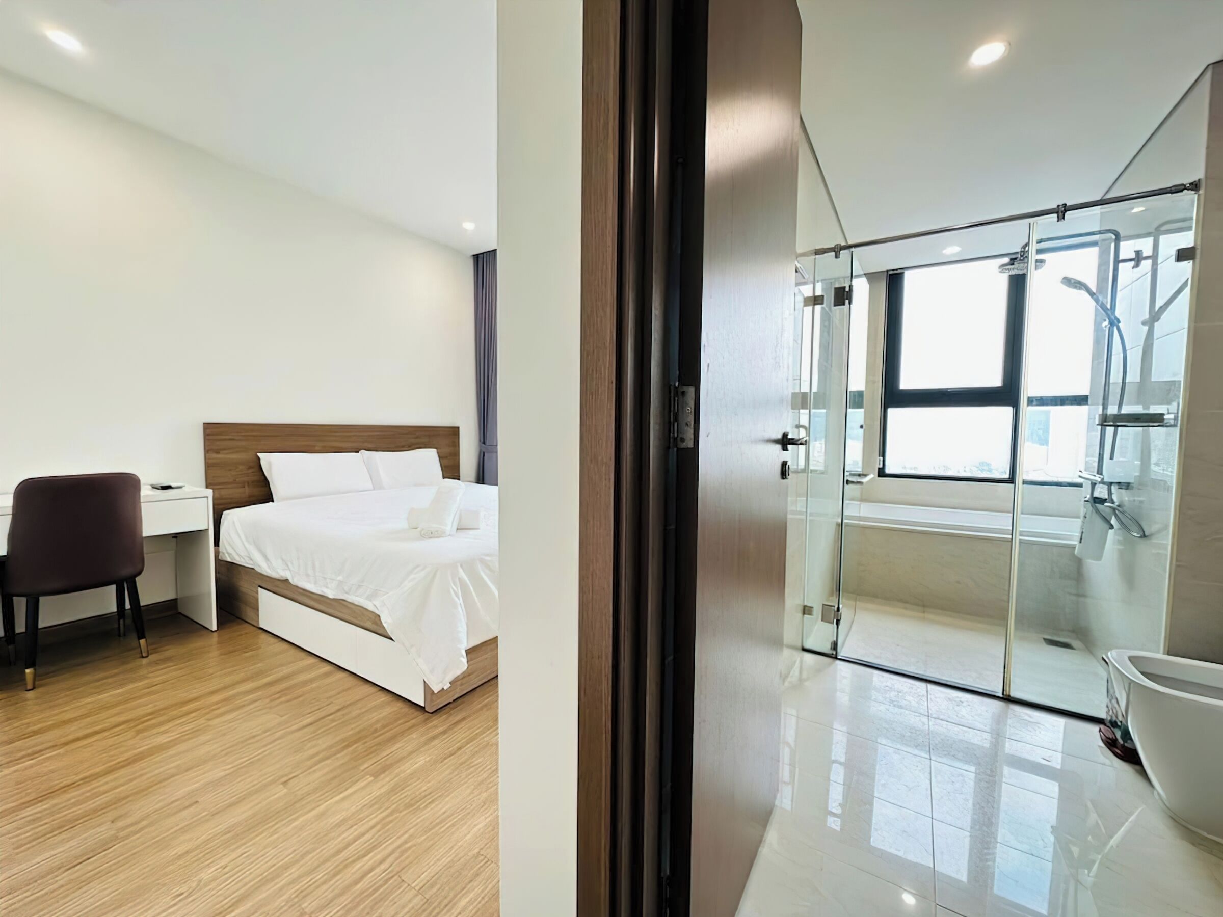 A modern hotel room with a bed, desk, chair, and a glass-enclosed shower area.