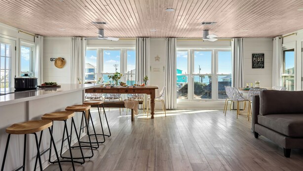 A modern kitchen with wooden flooring, a dining area with bar stools, and large windows offering a view of the outdoors. - Private Pool 20Steps2Beach Arcade Ping Pong Sleeps18 Barrel Sauna  (Gulf Shores)