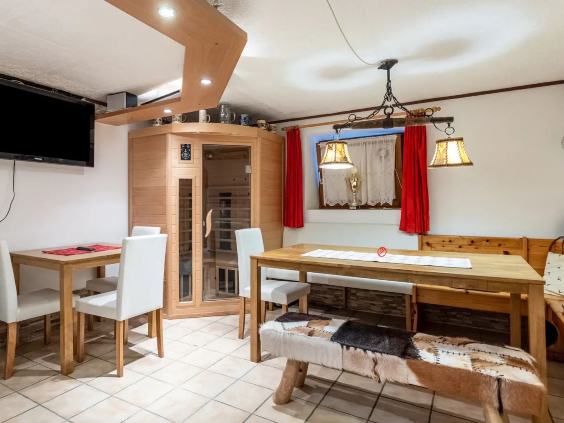 A dining area with a wooden table and chairs, a bench, a television mounted on the wall, and a window with red curtains.