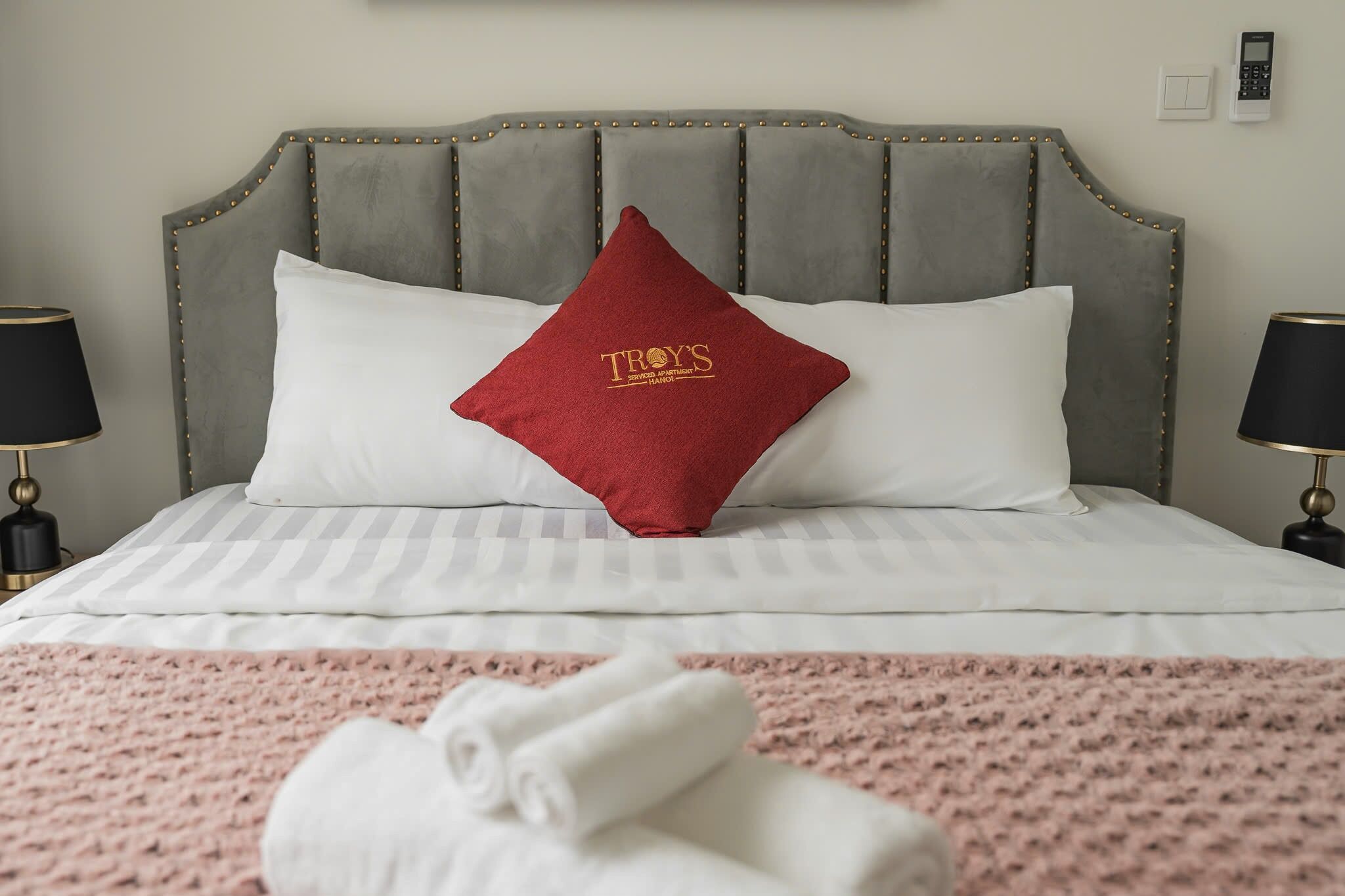 A neatly made bed with a grey upholstered headboard, a red pillow with "TROY'S" logo, and a pink textured blanket.