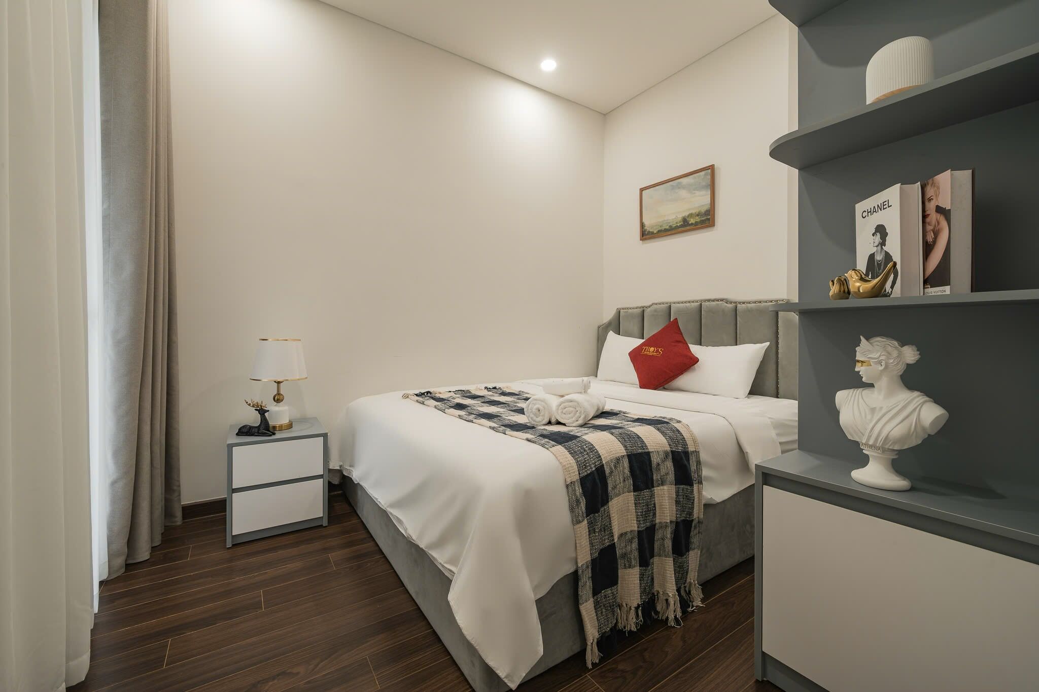 Un dormitorio ordenado con cama, mesita de noche, lámpara y un busto en una estantería.