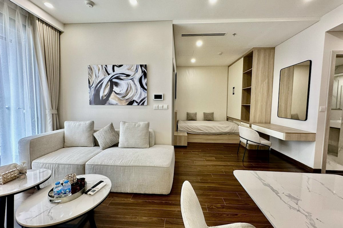 A modern hotel room with a sofa, a small table with water bottles, a bed, a chair, and a large mirror.