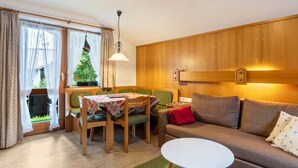 A cozy living room with a wooden wall, a sofa, a dining table with chairs, and a view of a green garden through a window.