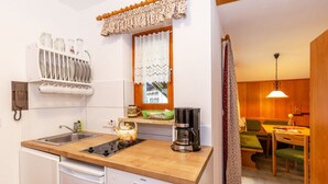 A compact kitchen with a sink, stove, and coffee maker. There is a window with curtains, a dining area with a table and chairs, and a wooden cabinet.
