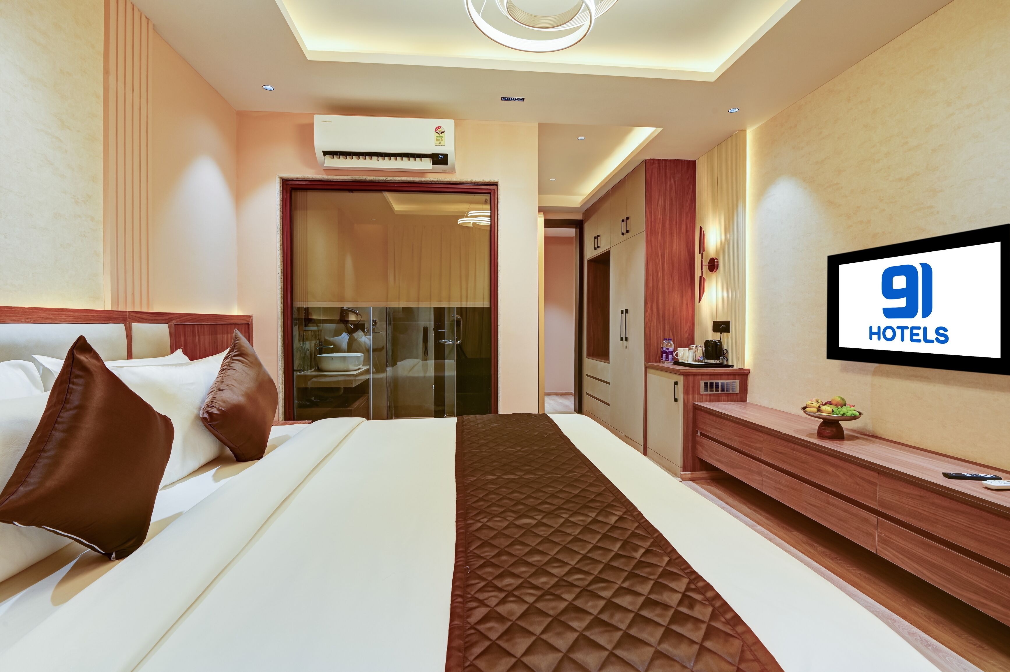 A hotel room with a bed, a TV, a minibar, and a bathroom visible through a glass door.